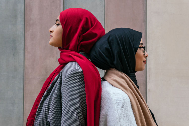 Side view of arab female friends in traditional hijab standing back to back on street and looking away