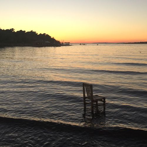 Abandoned empty chair on shore against sky during sunset