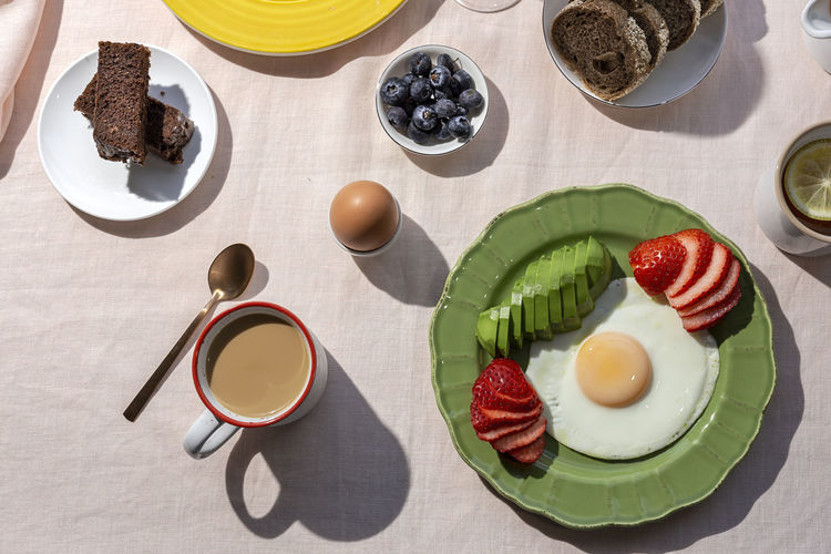 Homemade full healthy breakfast in sunlight with eggs, avocado, strawberries, blueberries, sponge cake, croissants, toast, tea, coffee and orange juice