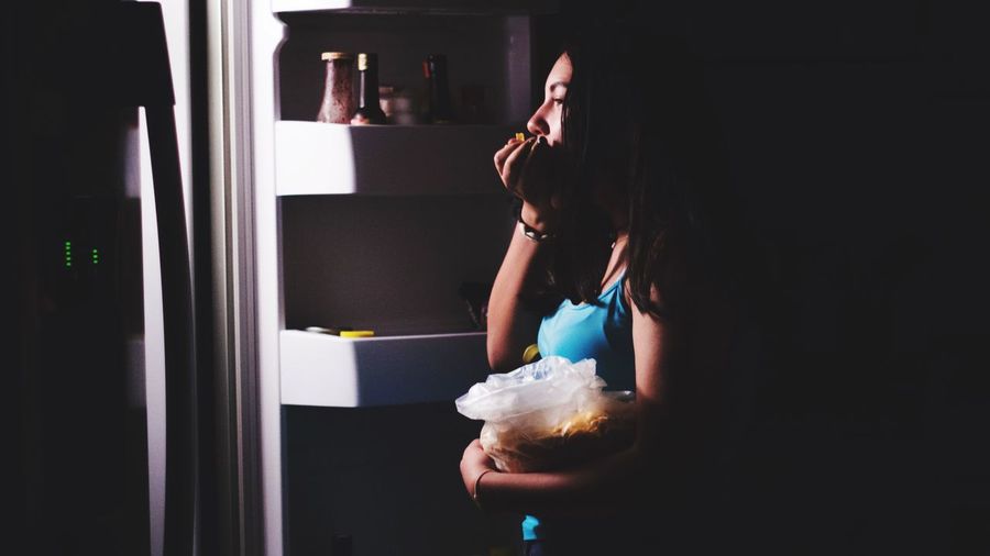 Side view of teenage girl eating while standing in dark at home