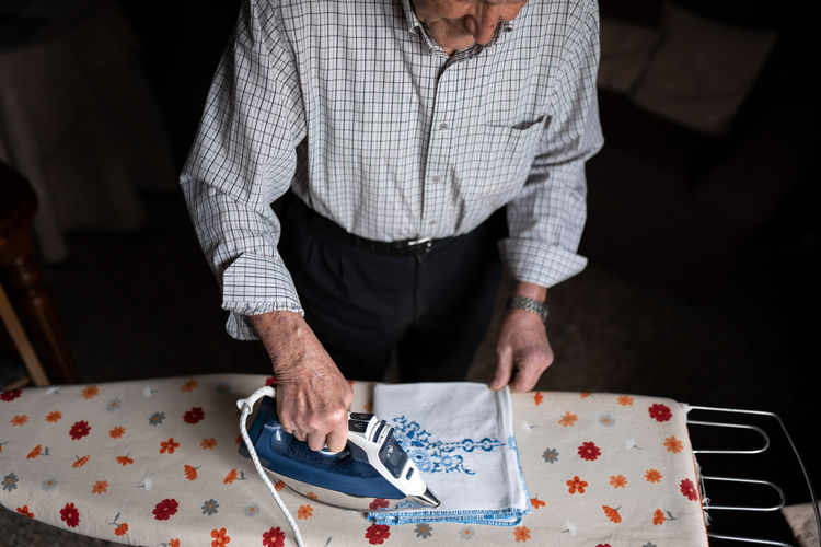 Cropped unrecognizable senior male in checkered shirt ironing fabric on board while standing in light room with couch at home