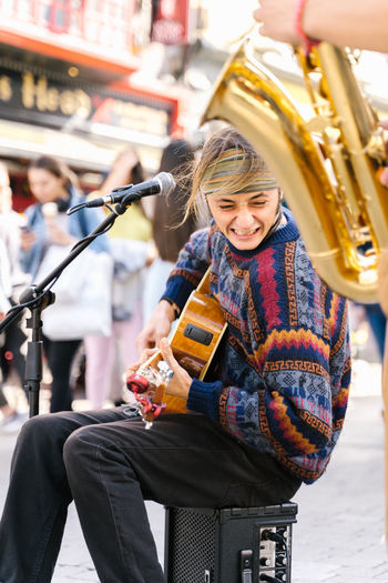 Vertical photo of a young man singing while playing guitar next to a saxophonist in the street