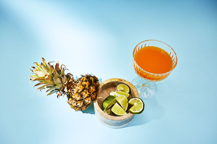 Directly above shot of orange drink on blue background with pineapple and lime garnishes
