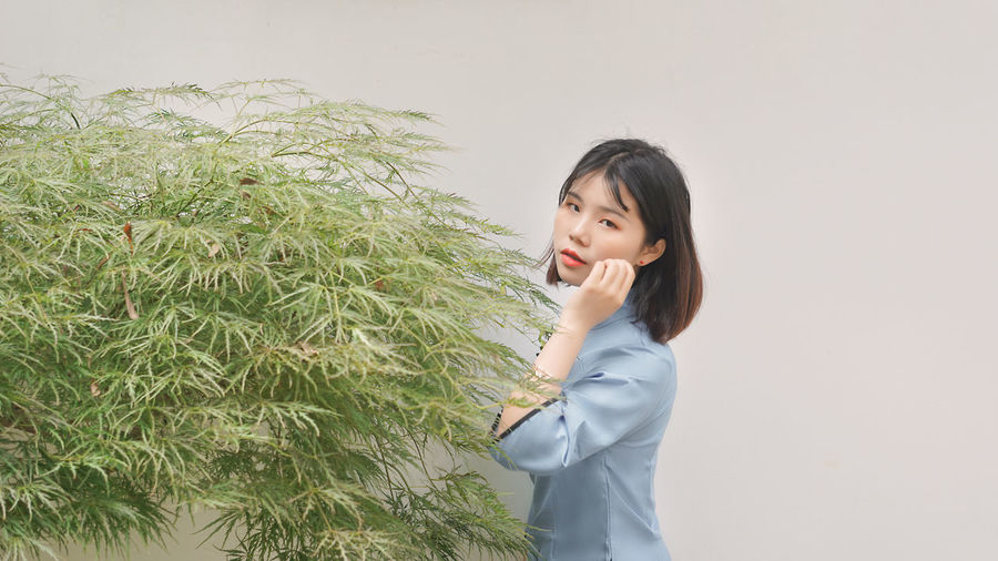 Portrait of beautiful young woman standing by plant against wall