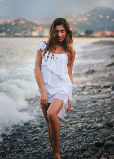 Portrait of young woman wearing white dress posing on sea shore at beach
