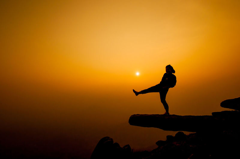 Silhouette woman kicking on cliff against sky during sunset