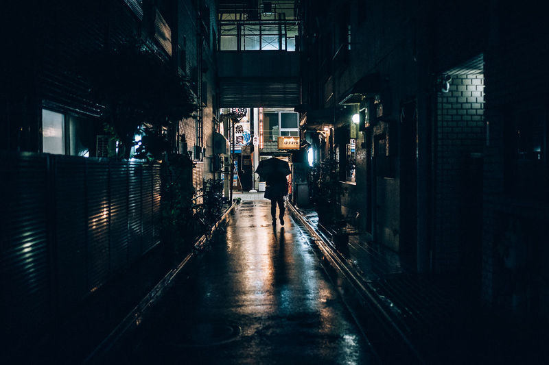 Rear view of man walking on illuminated alley amidst buildings at night