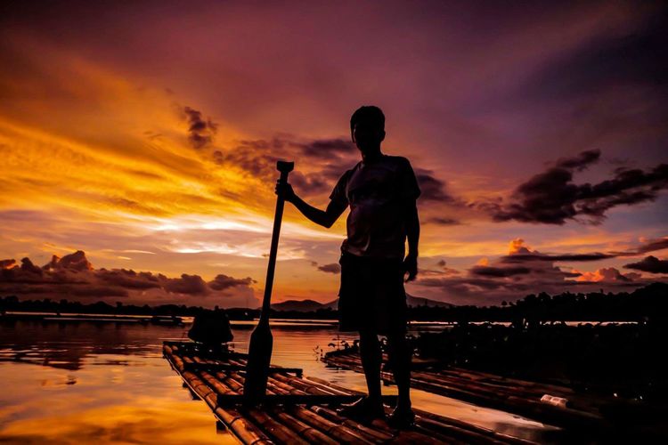 Man standing on wooden raft in lake against sky during sunset
