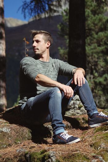 Young man looking away while sitting on rock in forest