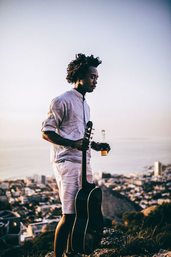 Mid adult man holding beer bottle and guitar while standing on rock against clear sky during sunset