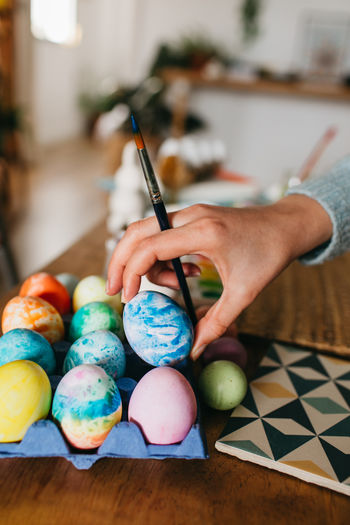 Unrecognizable person with brush arranging colorful eggs on carton on table while preparing for easter celebration at home person