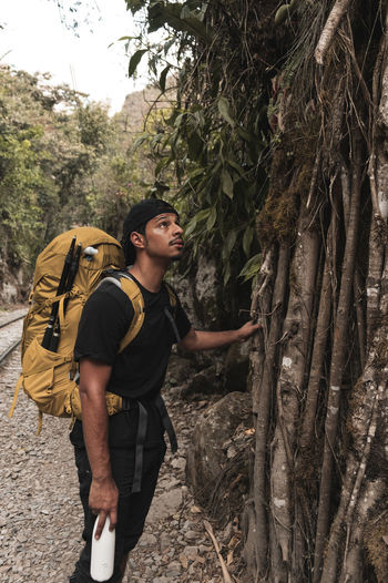 Side view of young man with backpack standing by tree in forest