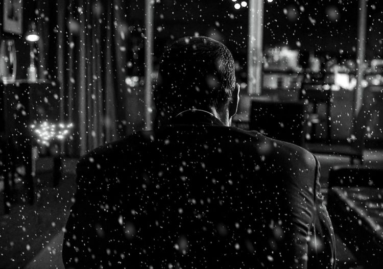 Rear view of man sitting at home seen from glass window during snowfall