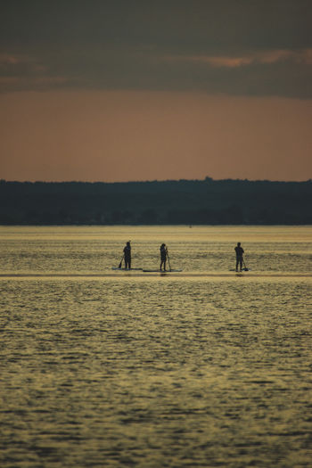 People paddleboarding in sea against sky during sunset