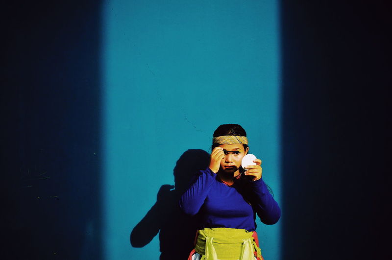Teenage girl applying make-up while standing against blue wall