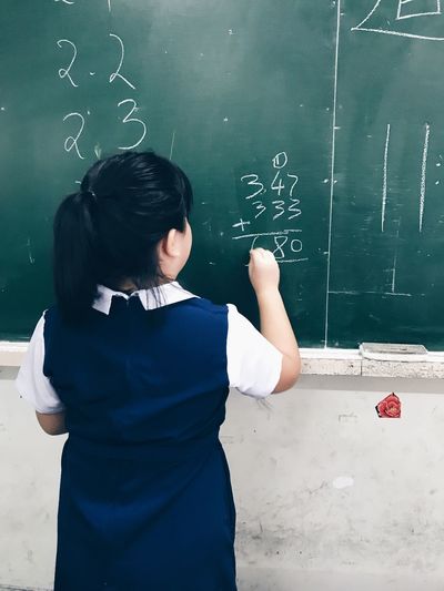 Rear view of girl writing on blackboard in classroom