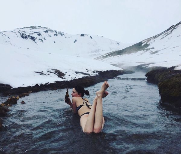 Young woman holding beer bottle while lying in stream amidst snowcapped mountain