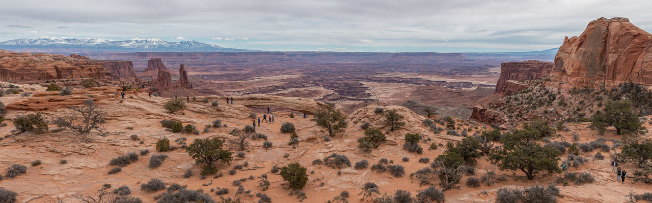 Panoramic view of tourists enjoying mesa arch with snow capped mountains in the background