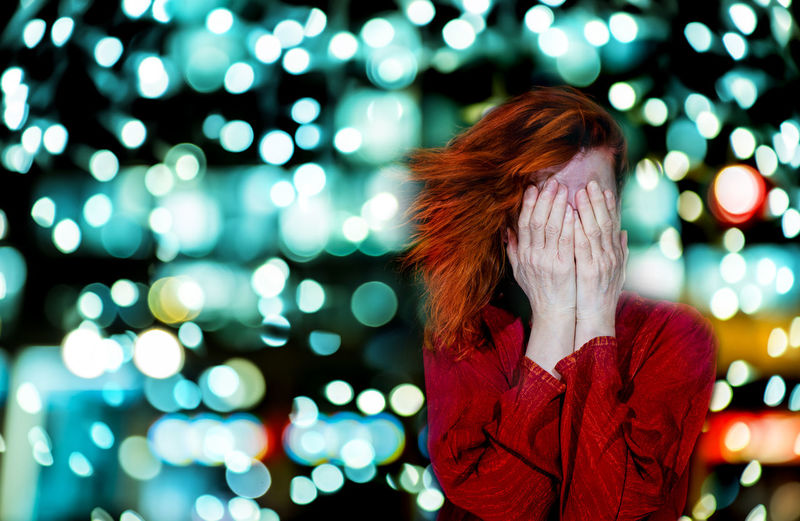 Woman with hands covering face standing against illuminated lights at night