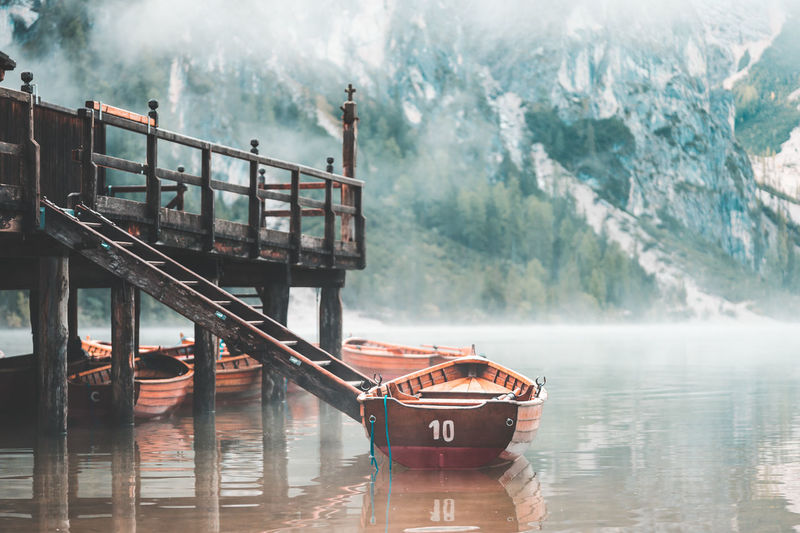 Boats moored by pier on lake against mountains during foggy weather