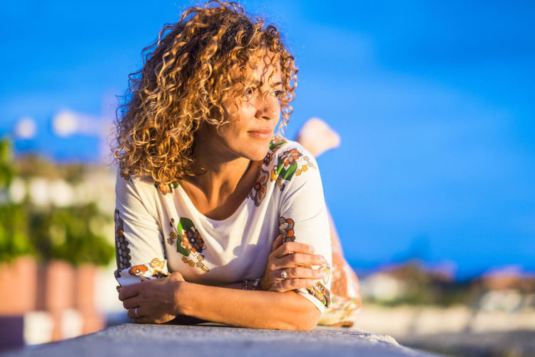 Woman with curly hair lying down on retaining wall against blue sky