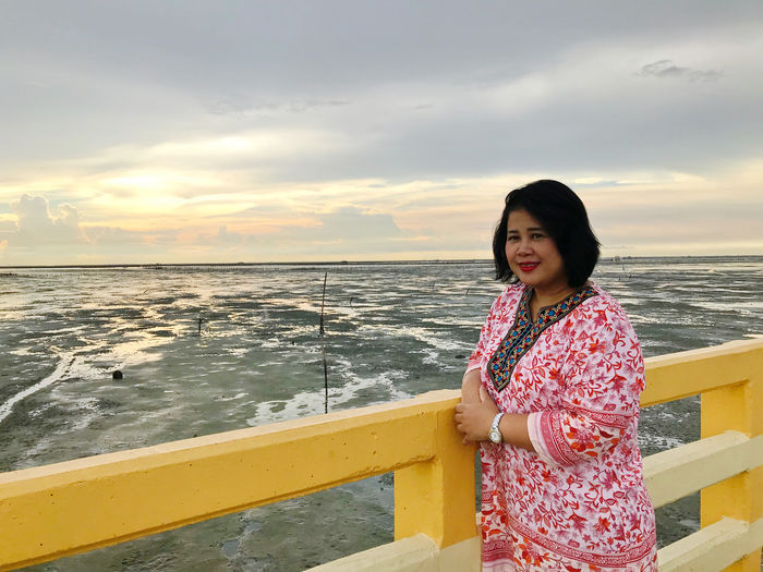Woman standing by railing against sea during sunset