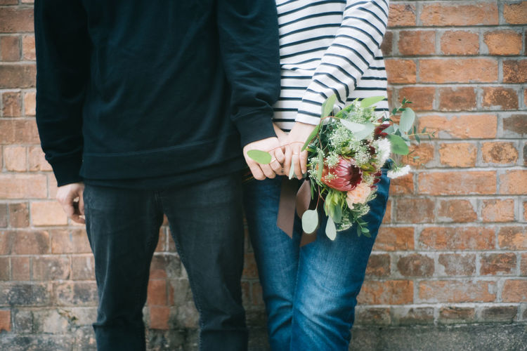 Midsection of couple with bouquet standing against brick wall