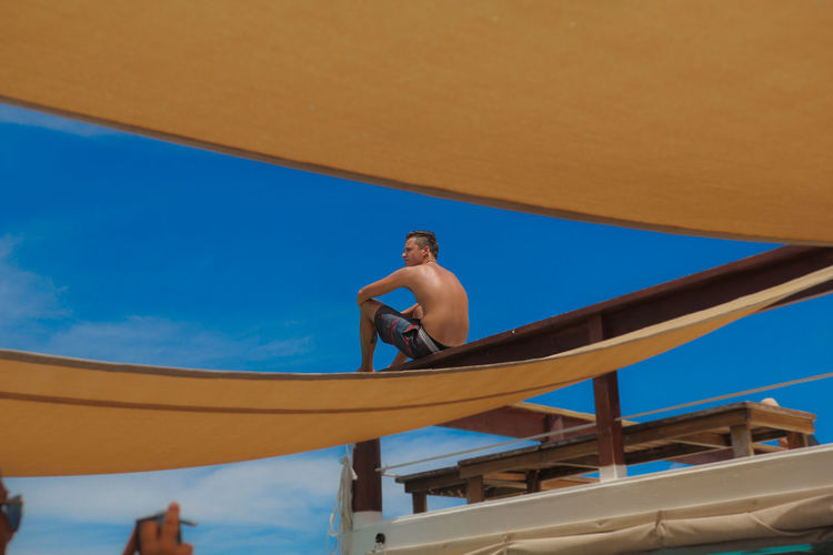 Low angle view of shirtless man sitting on pier against blue sky