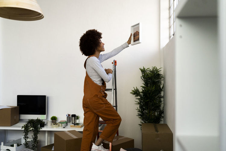 Afro woman putting picture frame on wall while climbing on ladder in new apartment