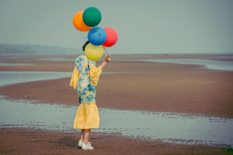 Rear view full length of young woman holding colorful balloons at beach