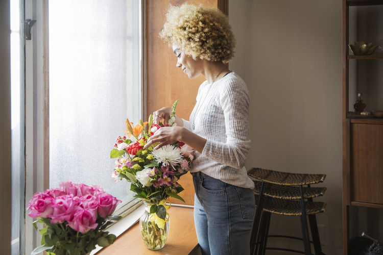 Smiling young woman arranging flowers in container on window sill at home