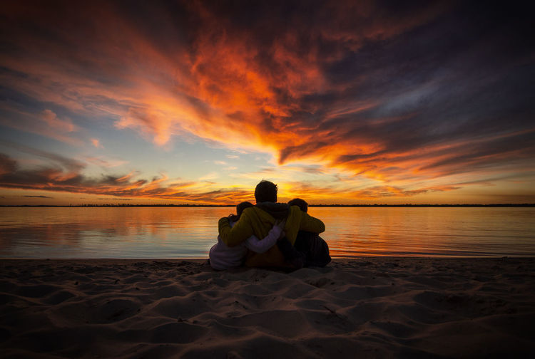 Rear view of people embracing each other while looking at lake against dramatic sky during sunset