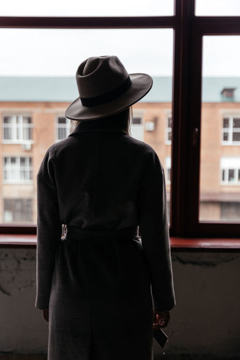 Silhouette of a woman in a beige autumn coat and hat on the background of the window