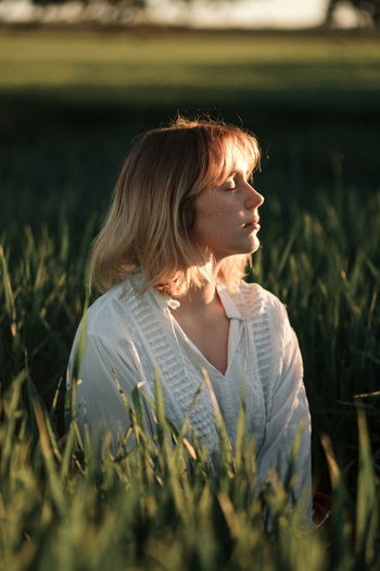 Peaceful young female in retro styled white blouse sitting amidst tall green grass and closed eyes while resting in summer evening in countryside