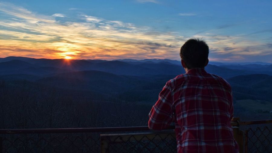Rear view of man standing at railing against mountains during sunset