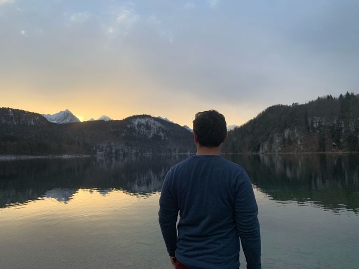 Rear view of man looking at lake against cloudy sky during sunset