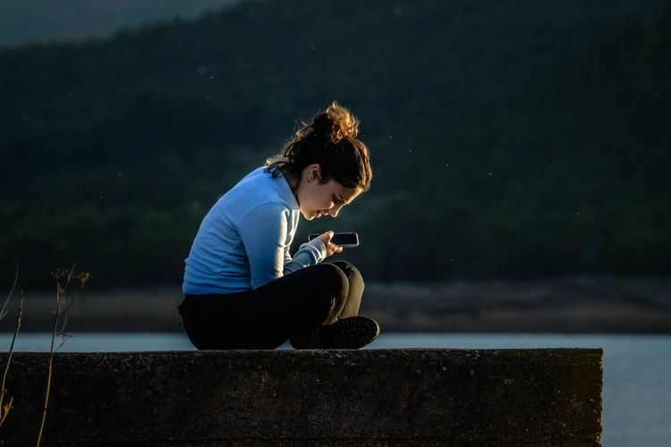 Girl using mobile phone while sitting on retaining wall by lake