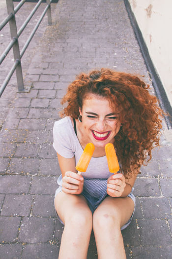 High angle view of cheerful woman having orange popsicles on footpath
