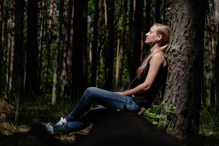 A girl in a forest sits leaning against a tree, headphones listening to music. positive emotion.