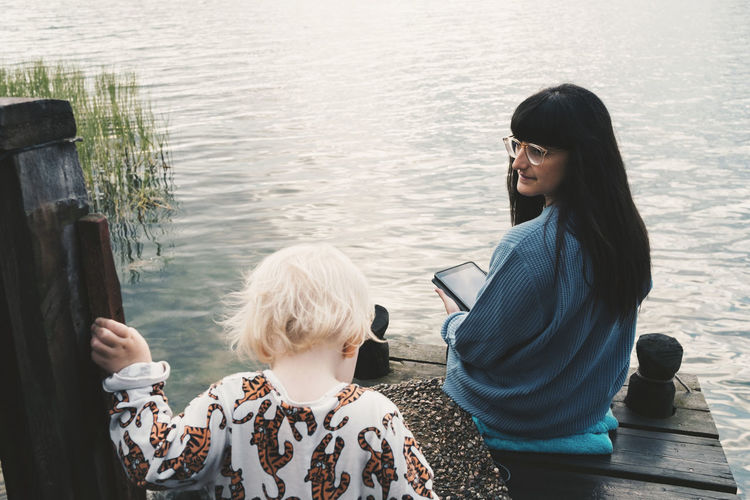 Mother looking at daughter while holding digital tablet and sitting on jetty over lake