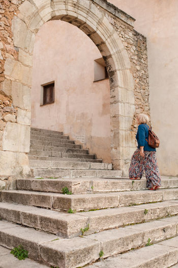Side view of female tourist strolling up stone steps of medieval building with arched passage during sightseeing in cuenca town in spain