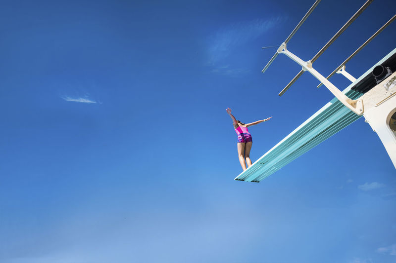 Low angle view of carefree girl standing on diving platform against blue sky during sunny day