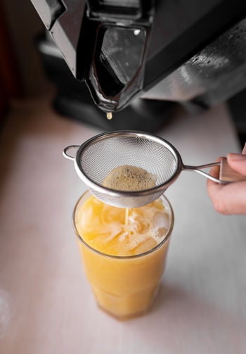 From above of crop anonymous person straining tasty fruit drink through sieve into glass placed on table in light kitchen person