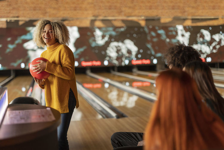 Smiling young woman holding ball with friends sitting at bowling alley