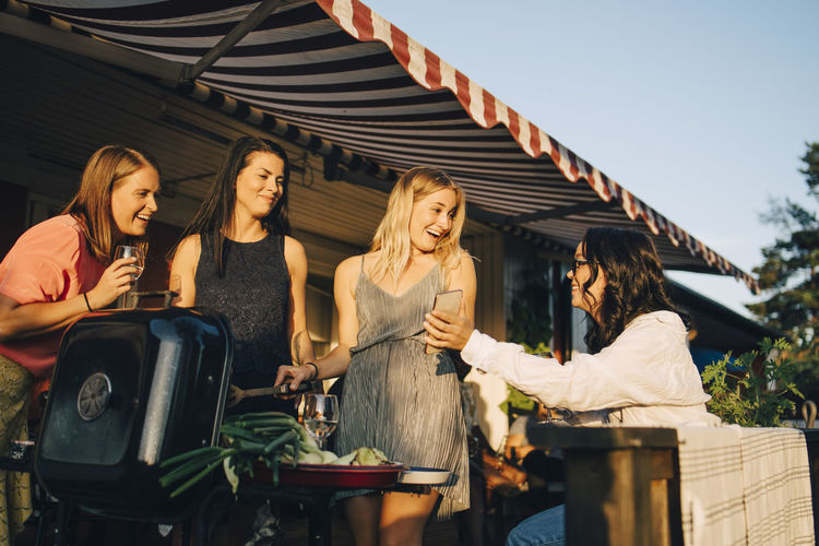Smiling woman showing mobile phone to friends grilling food on barbecue