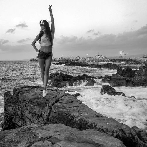 Young woman standing with arms raised on rock by sea against sky