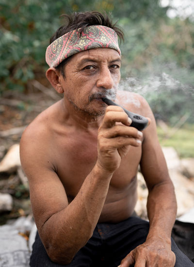 Serious bearded shirtless mexican male in bandana sitting on ground and looking at camera while smoking pipe  person