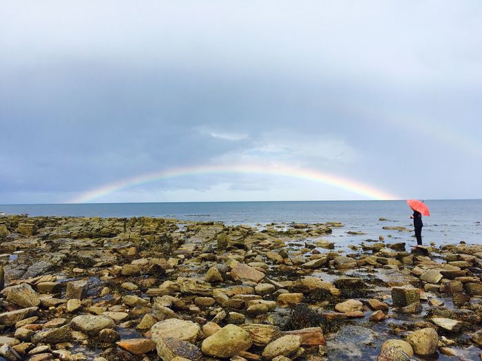 Person standing by sea with rainbow against sky during rain