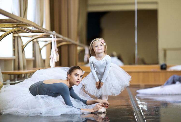 An adult ballerina is tying pointe shoes to a little ballerina.
