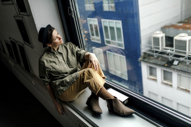 Fashionable woman looking through window while sitting on window sill at studio